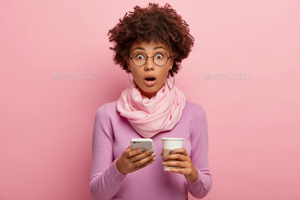 Stupefied emotional dark skinned woman has curly Afro hair, checks email on smartphone device, drink