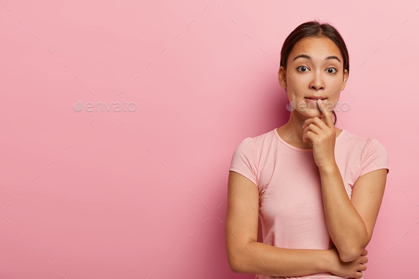 Photo of confused Asian girl bites lips and looks nervousy at camera, holds hands partly crossed, we