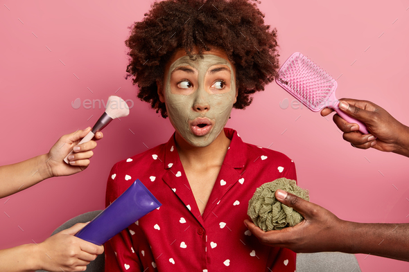 Spa, health care concept. Surprised black young woman with clay mask, looks aside, dressed in domest