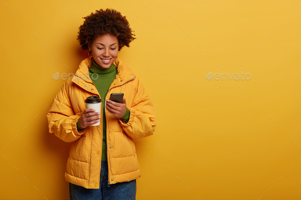 Photo of attractive positive woman in yellow outerwear, happy to read good comments under post, hold
