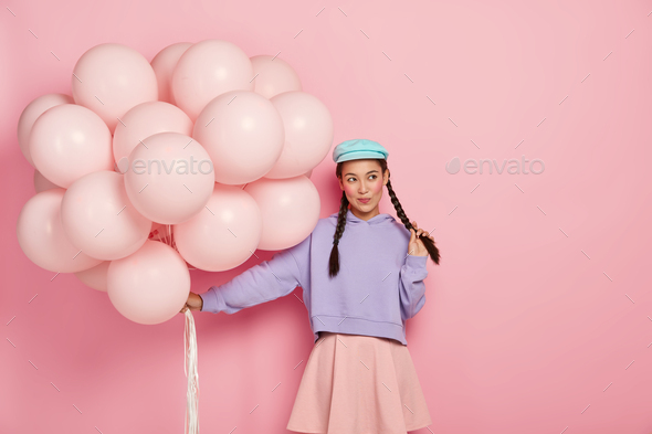 Pensive pretty girl with rouge cheeks, dressed in fashionable clothes, holds bunch of balloons, come