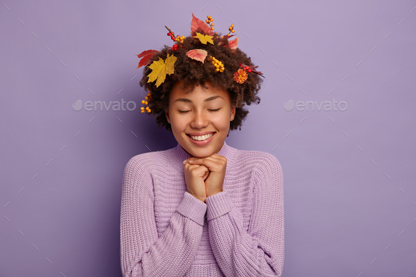 Indoor portrait of lovely happy woman keeps both hands under chin, enjoys warm dry autumnal weather,