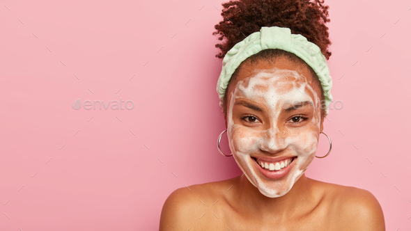 Pretty overjoyed dark skinned lady uses facial foam for washing cosmetic off on face, has broad smil