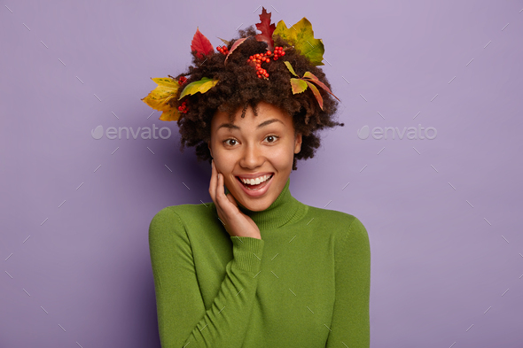 Portrait of lovely cheerful Afro woman keeps hand on cheek, wears green turtleneck, has foliage in d