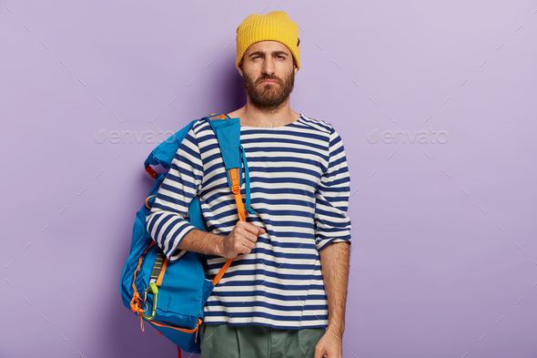 Displeased unshaven man smirks face, has unhappy expression, carries tourist rucksack, being tired o