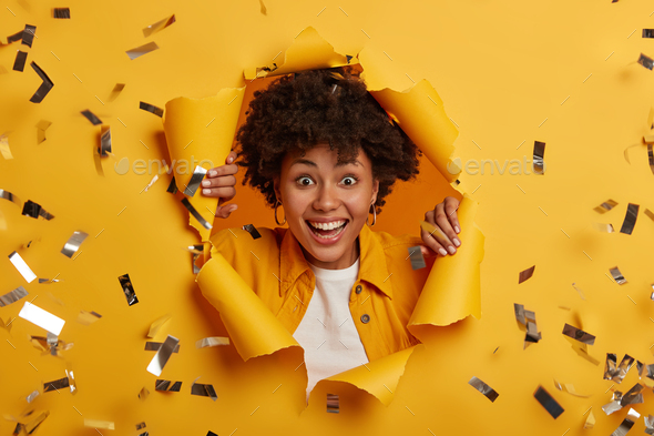 Close up shot of overjoyed Afro American female stands in ripped hole of yellow background, dressed