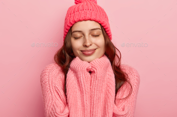 Headshot of satisfied European woman has healthy skin, keeps eyes shut, hands under chin, wears warm