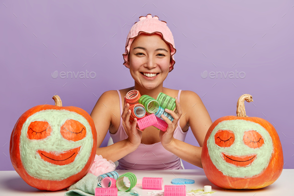 Photo of happy mixed race woman wears bath cap, holds multicolored curlers, has hygienic procedures