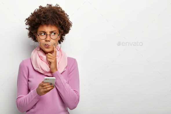 Pensive dark skinned woman looks thoughtfully aside, holds mobile phone, waits for call, purses lips
