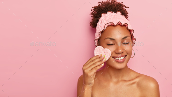 Photo of relaxed dark skinned female model wipes face with cosmetic sponge, removes makeup, wears pi