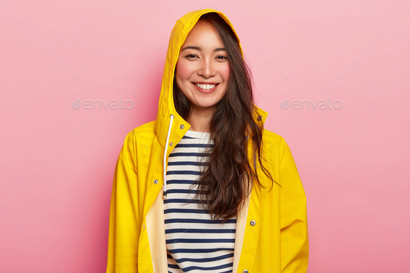 Smiling beautiful woman enjoys wearing warm striped jumper, yellow raincoat with hood, has good mood