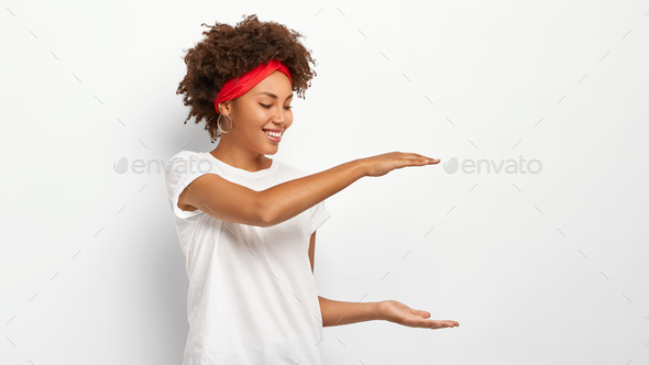 Horizontal shot of happy dark skinned curly haired woman shows height of something, gestures with bo