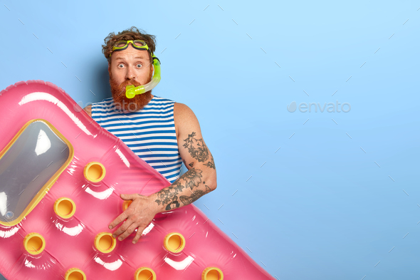 Curly ginger haired man wears swimming goggles, snorkeling mask and inflated pink mattress, ready fo