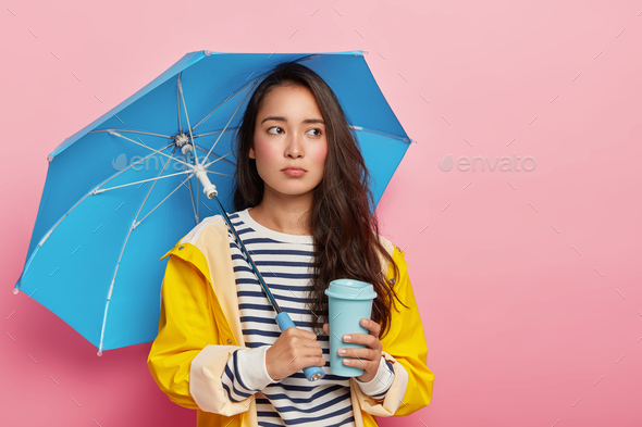 Sad woman feels depressed during cloudy rainy day, has seasonal depression, poses under waterproof u