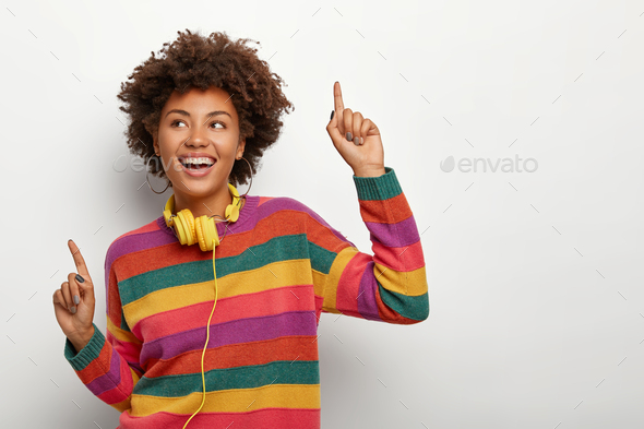 Cheerful Afro American woman raises arms and points with index fingers, dances happily to music, wea