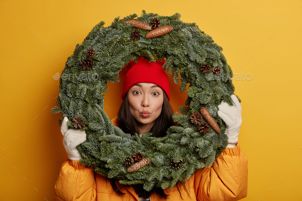 Adorable young Korean woman keeps lips rounded, looks through green spruce wreath, wears yellow coat