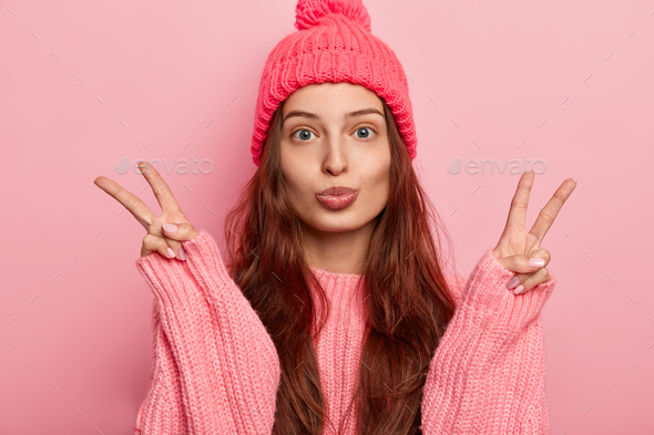Young beautiful brunette European woman keeps lips rounded, makes victory peace gesture, wears knitt