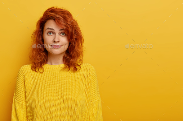 Beautiful young ginger woman wears bright knitted yellow sweater, raises eyebrows with wonder, looks