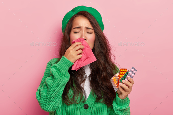 Unhappy brunette girl suffers from flu symptoms, rubs nose with handkerchief, has cold, holds pills,