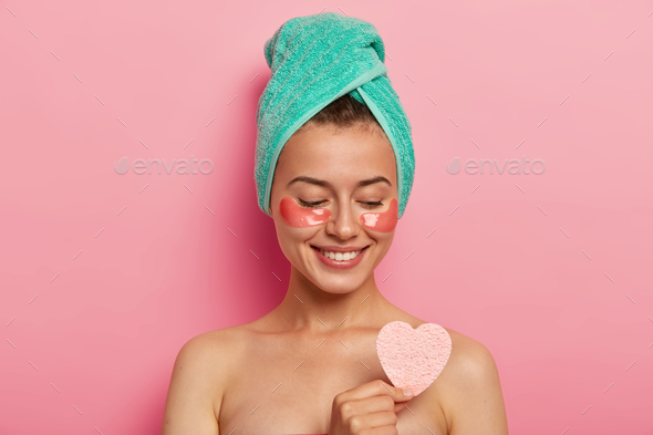 Cute European lady enjoys eye skin treatment, holds cosmetic sponge over naked body, has gentle smil