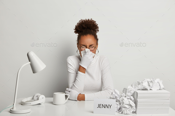 Photo of ill office worker has sneezing and running nose, symptoms of flu, wears white turtleneck an
