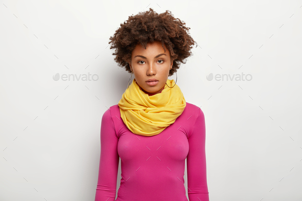 Half length shot of serious dark skinned curly haired woman wears big round earrings, yellow scarf a