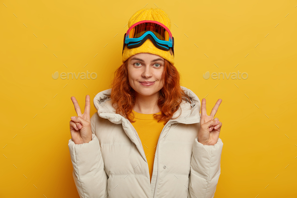 Half length shot of ginger pretty woman raises arms, makes peace or victory gesture, uses ski goggle