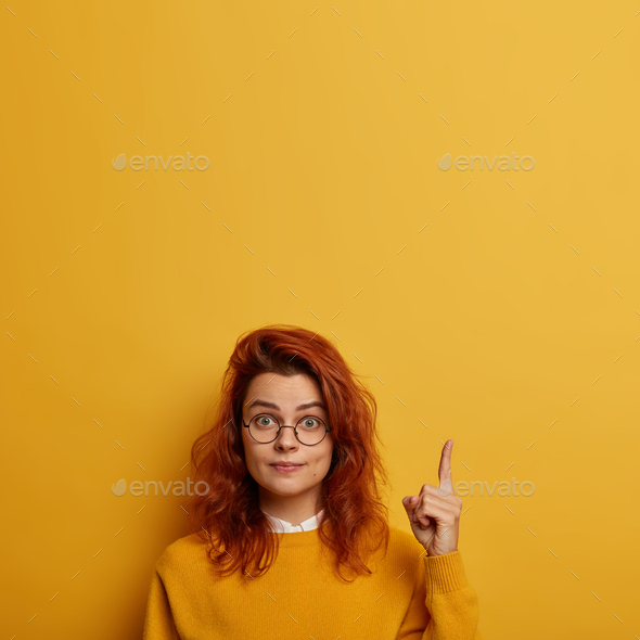 You need look. Surprised redhead European woman wears round glasses, points above, shows brand new p