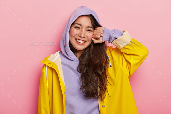 Positive brunette Korean girl in casual hoody and raincoat, smiles pleasantly, being in good mood af