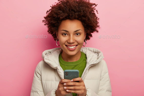 Portrait of glad dark skinned hipster girl uses mobile phone for chatting with followers, checks ema
