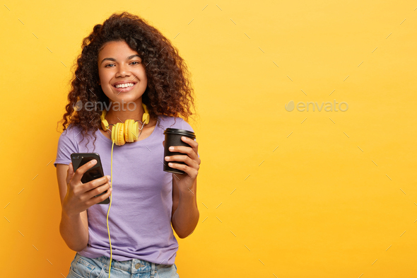 Smiling curly haired female watches video on mobile phone during coffee break, listens audio tracks
