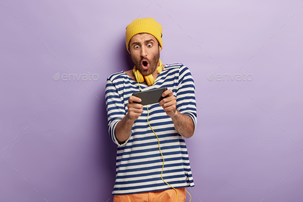 Photo of shocked unshaven guy holds mobile phone horizontally, holds breath, plays games online, bei