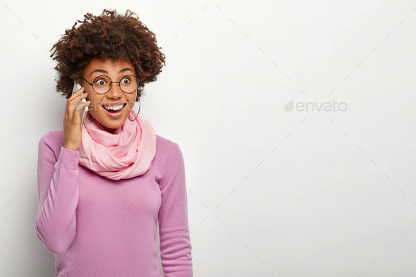 Horizontal shot of cheerful young African American lady calls someone via modern cellphone, uses mod