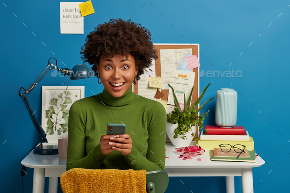 Photo of happy Afro American female student poses with smart phone, spends time for course work, sit