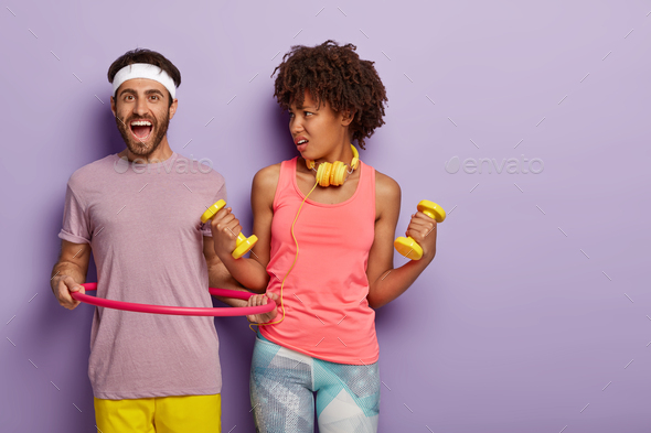 Mixed race people have workout, hold dumbbells in both hands, man exercises with hula hoop, dressed