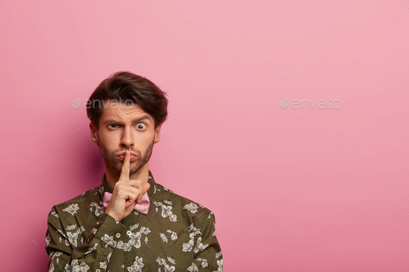 Serious stylish man makes silence gesture, gossips or tells secret, holds index finger over lips, as