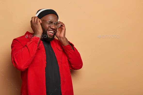 Plump black man stands half turned against beige wall, listens audio track, enjoys loud sound in hea