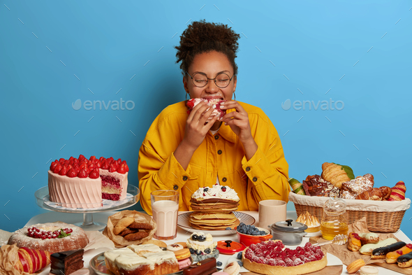 Cheat meal and gluttony concept. Ethnic curly woman eats strawberry creamy cake with much calories,