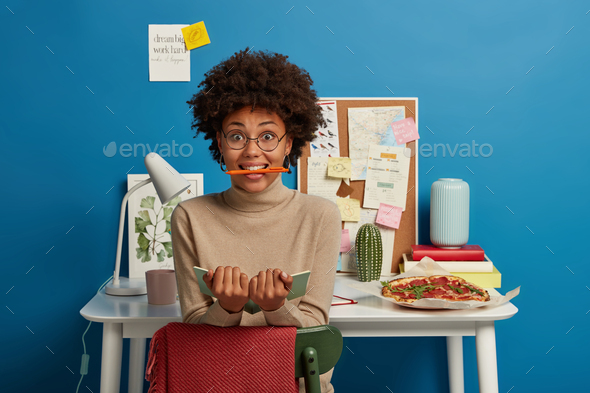 Surprised Afro American woman has fun while working, keeps pen in mouth, holds opened notebook for w