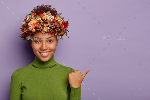Photo of dark skinned young curly woman points thumb away, wears green turtleneck, autumnal wreath,