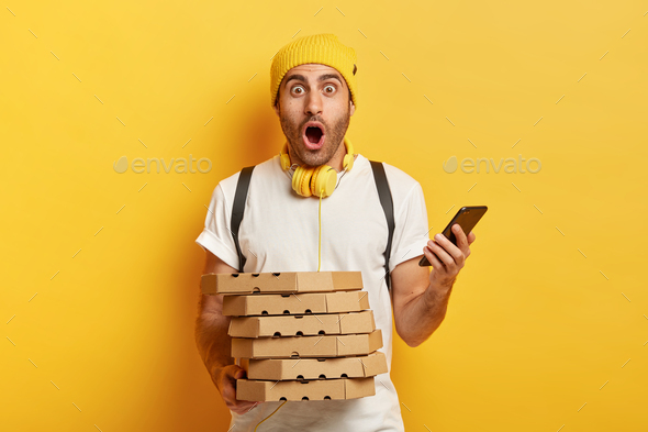 Amazed deliveryman receives orders from customers via smartphone, holds pile of cardboard pizza boxe