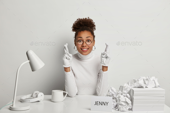 Photo of lovely curly woman crosses fingers, wishes good luck, sits at desktop, hopes for successful