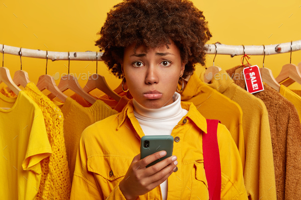 Dejected Afro woman uses smartphone for online shopping, unhappy cannot find schedule of sales, stan