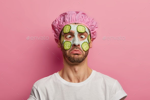 Young man crosses eyes, applies cream mask on face with cucumber, has beauty session, wears bath cap