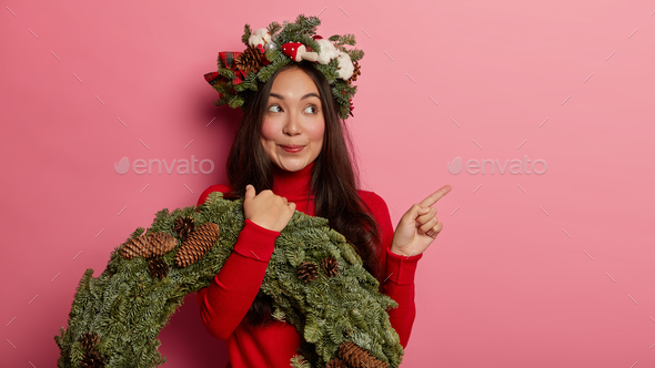 Photo of lovely young Asian woman points index finger holds spruce wreath, wears casual red turtlene