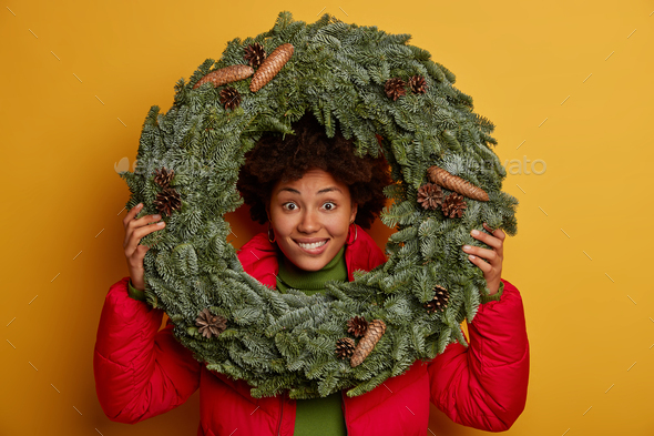 Close up shot of happy dark skinned woman looks through round spruce wreath, dressed in red jacket,