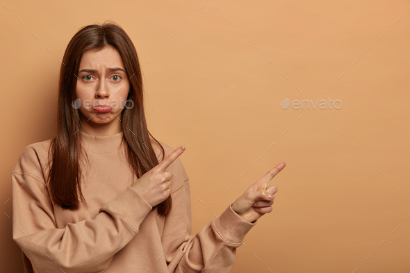 Indoor shot of unhappy disappointed woman points on blank space, missed opportunity, asks your opini