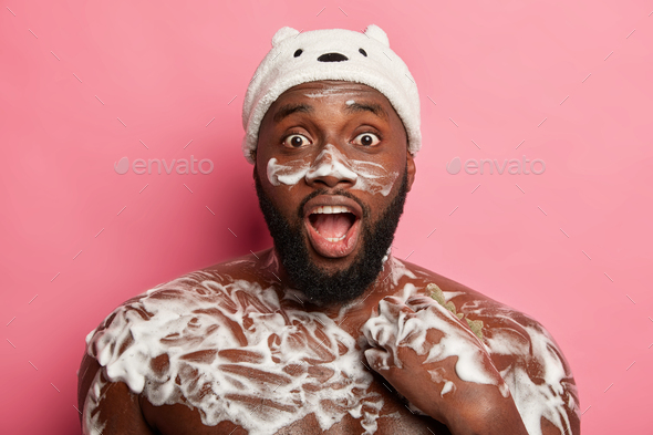 Healthcare, hygiene, spa concept. Shocked Afro man washes body with soap sponge in douche, feels fre
