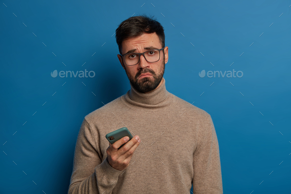 Displeased bearded guy smirks face, uses modern cellphone, cannot download application, has sad expr