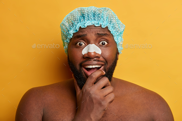 Black surprised bearded man holds chin, wears clear up strip on nose to remove blackheads, wears bat